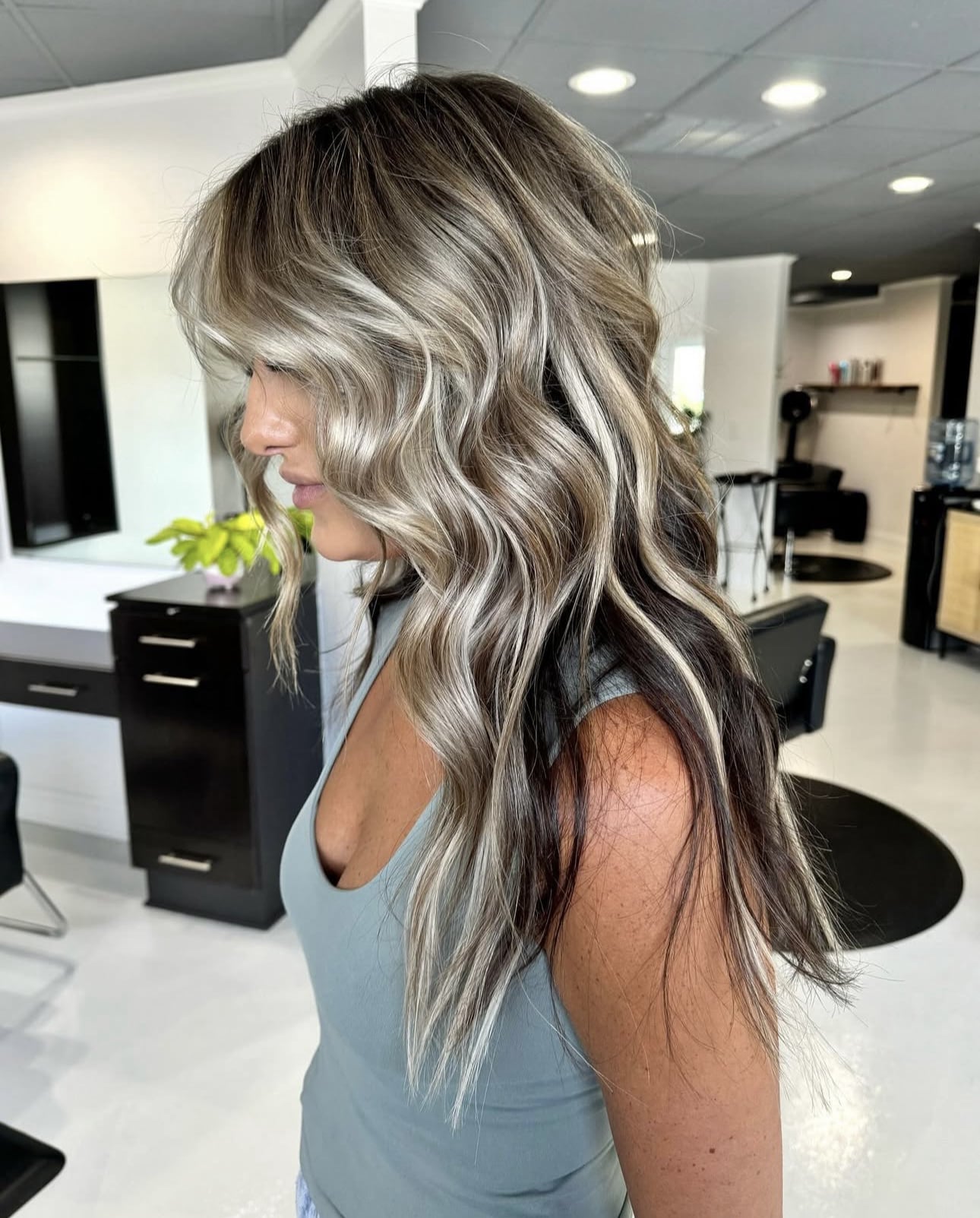 Side profile of a woman with long, wavy blonde highlights and dark lowlights in a hair salon setting