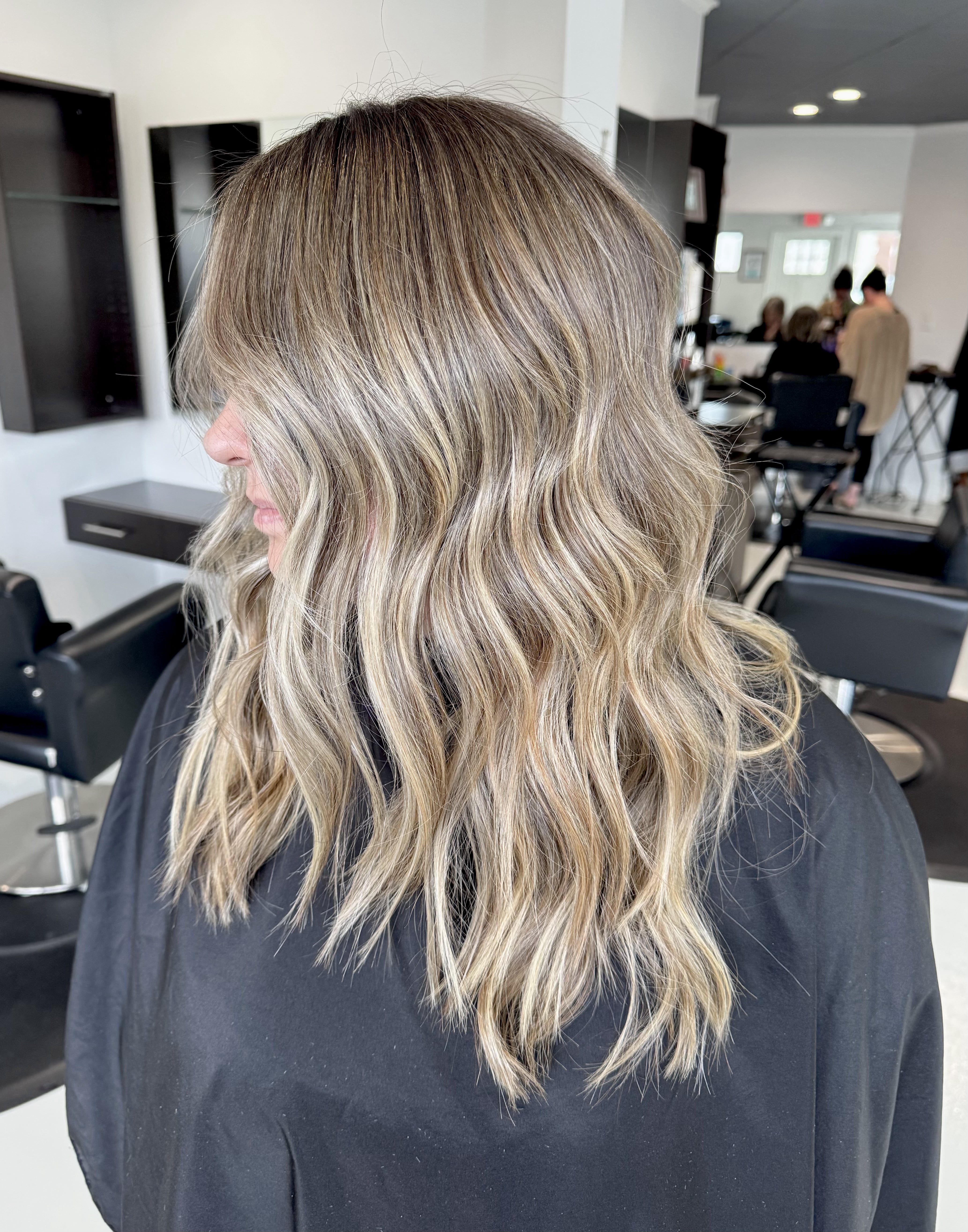 Side profile of a woman with long, wavy blonde balayage hair sitting in a salon chair
