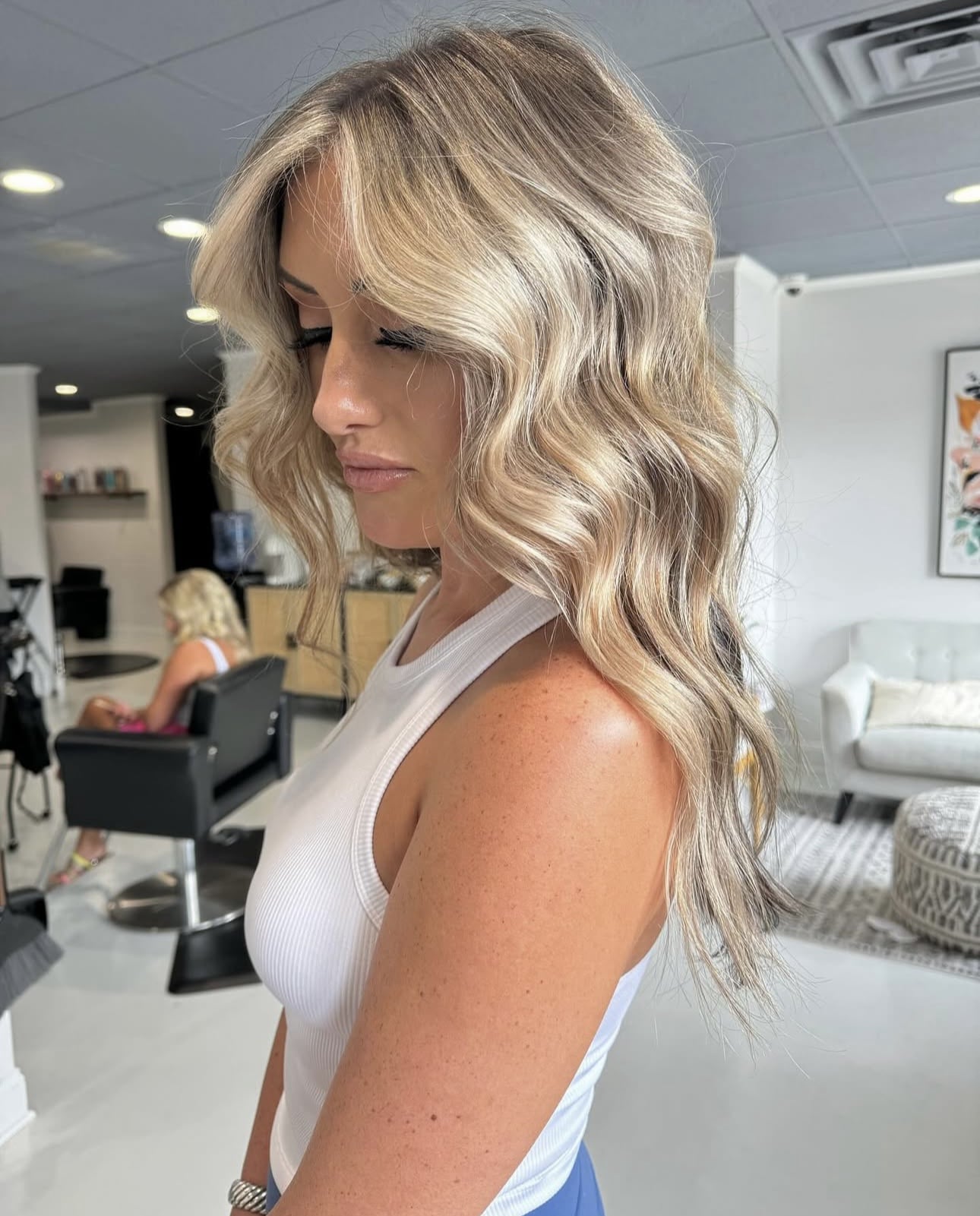 Side profile of a woman with long, wavy blonde balayage hair and a white tank top in a salon